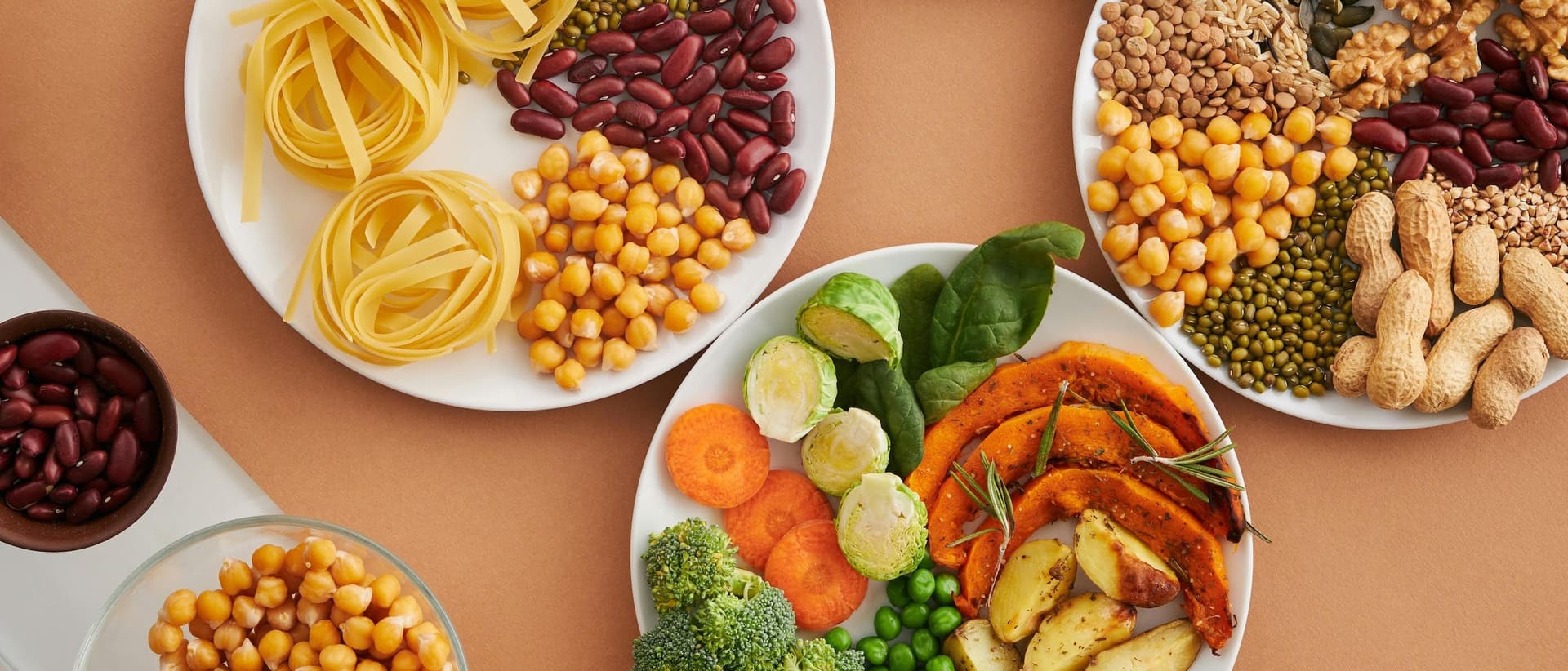 Multiple white plates filled with a diverse array of healthy grains, legumes, pasta, and roasted vegetables.